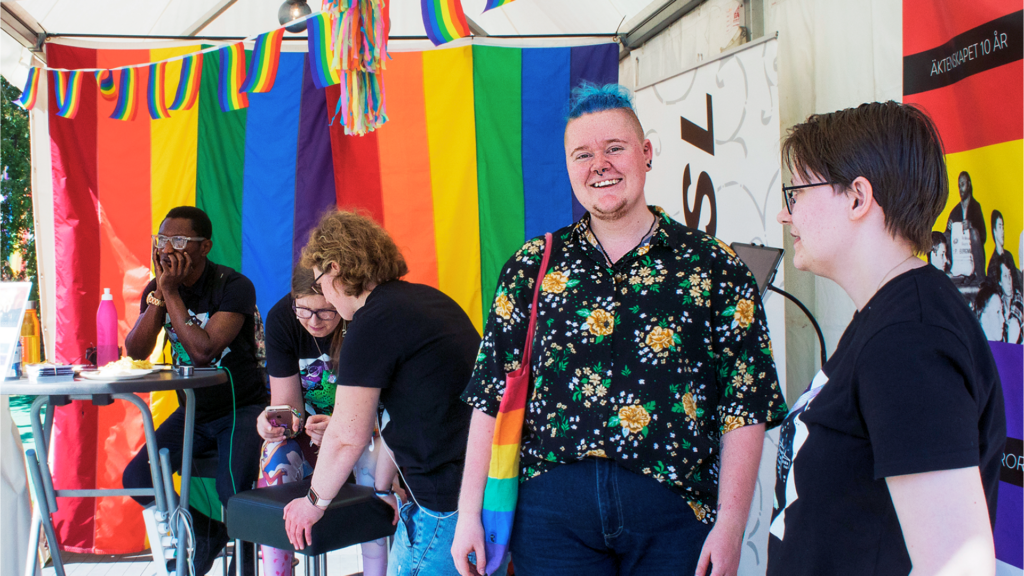 Foto på fem personer i vårt tält från EuroPride i Göteborg 2018. Två av personer står upp, resten sitter vid ett bord i bakgrunden. I taket hänger flera regnbågsflaggor.