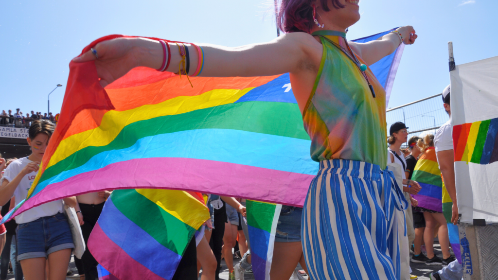 Foto på deltagare i vårt paradblock från Stockholm Pride 2019. I framgrunden går en person som håller upp regnbågsflaggan med båda armarna. I bakgrunden syns fler deltagare med regnbågsflaggor, åskådare på en bro och en klarblå himmel.