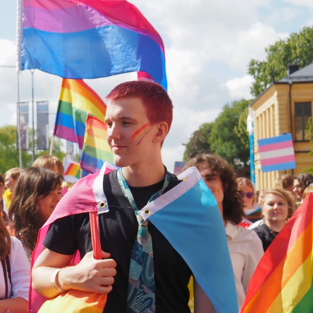 Foto på deltagare i vårt paradblock från Stockholm Pride 2023. En person är insvept i flaggan för pansexuella och har en sminkning och tygkassa med regnbågsflaggan. Den har svarta kläder och en mönstrad slips.