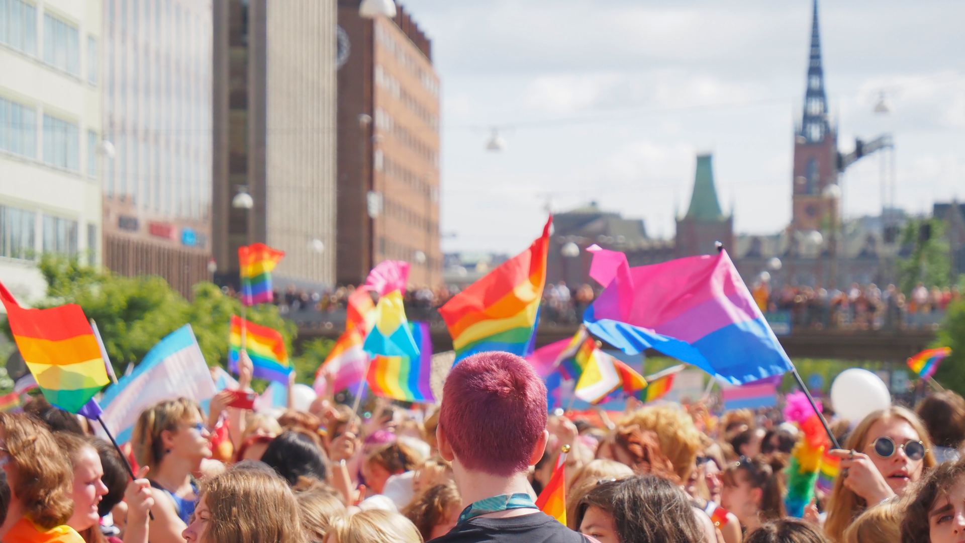 Foto på deltagare i vårt paradblock från Stockholm Pride 2023. Många viftar med regnbågsflaggor: några viftar med flaggan för bisexuella, transpersoner och lesbiska.