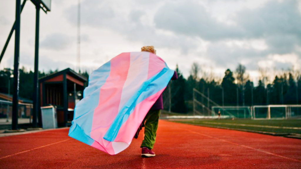 Foto på en person som springer på en löpbanan med en flaggan för transpersoner. Personen har lila tröja, gröna byxor och röda skor. I bakgrunden syns en grå himmel.