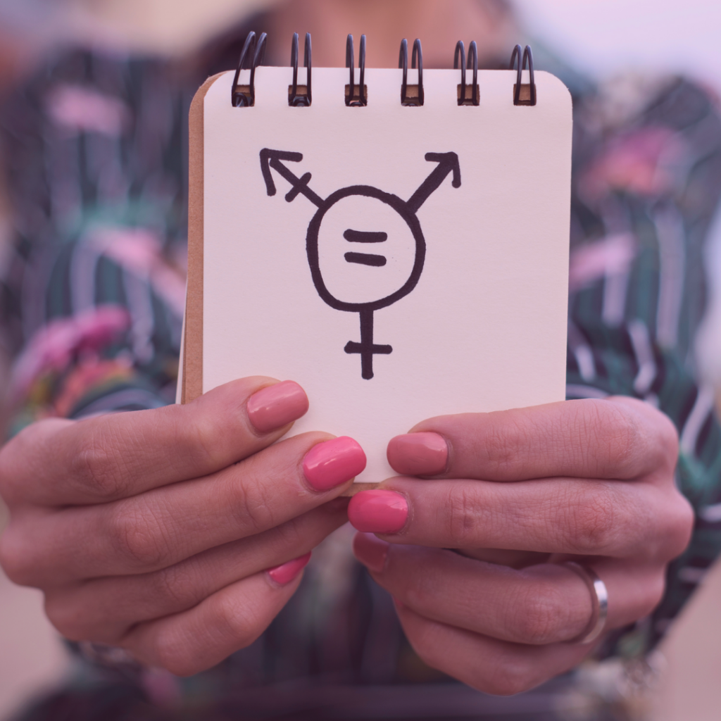 Foto på en anteckningsblock med en tecknad transsymbol som hålls upp an en person. Den har rosa naglar och vitrandig blues.
