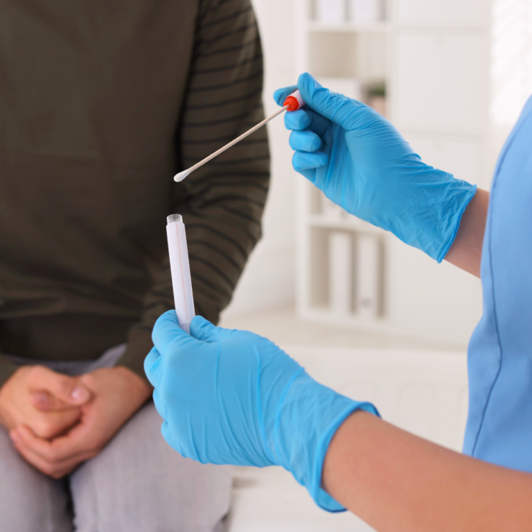 Närbild på en person som håller upp en bomullspinne och provrör. Personen har på sig ljusblåa engångshandskar och ljusblå kläder. I bakgrunden sitter en person på en brits. Den har en långärmad brun tröja och gråa byxor.