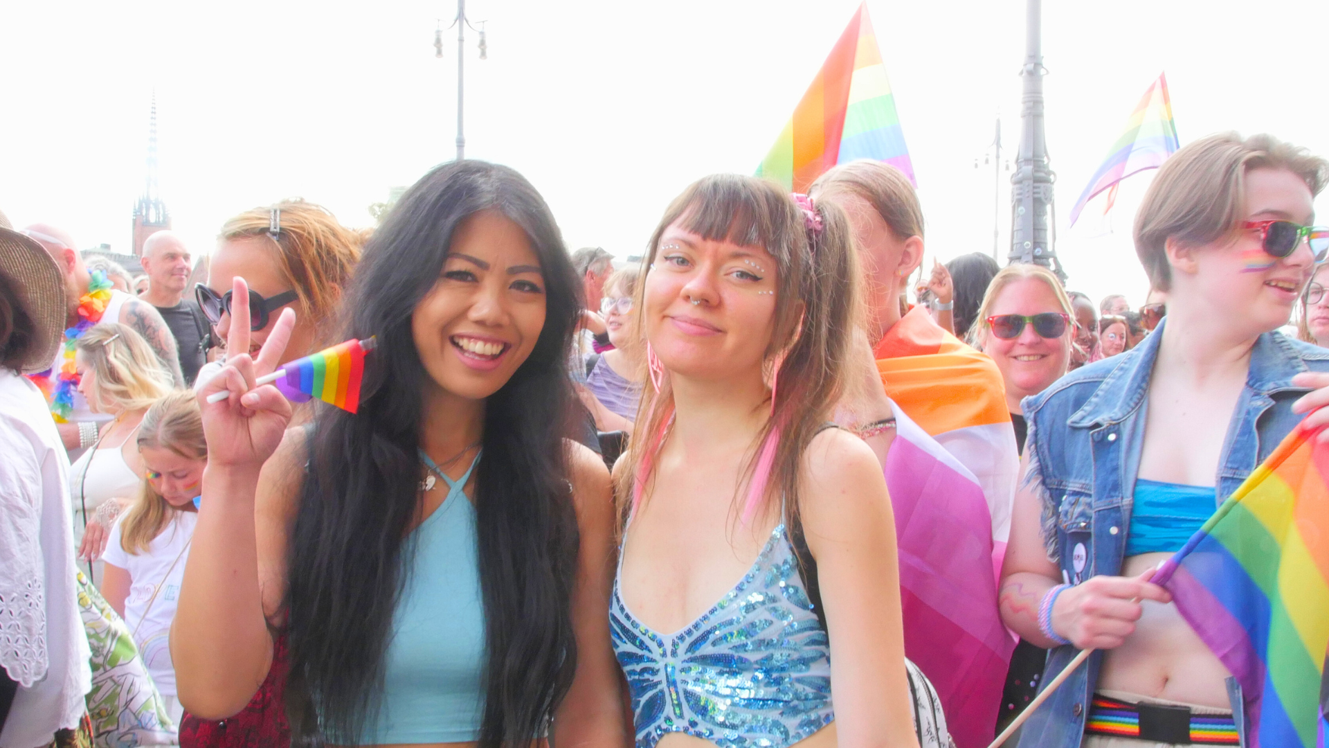 Foto på deltagare i vårt paradblock från Stockholm Pride 2024. Två personer håller hand och ler mot kameran: den ena håller i en liten regnbågsflagga och gör peace-tecknet.