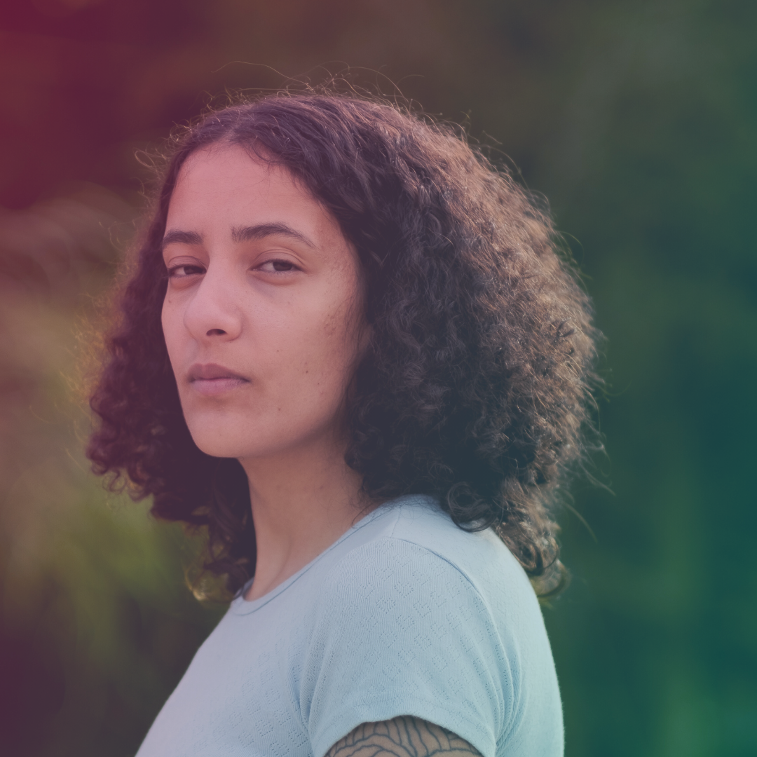 Foto på en person med krusigt hår och en grå t-shirt. Hen vänder ansiktet mot kameran och ser allvarlig ut.