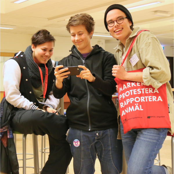 Foto på tre personer samlade runt en smartphone. Personerna har olika frisyrer, kläder och accessoarer.