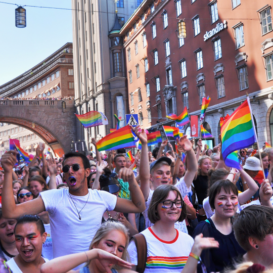 Foto på personer som går i en prideparad genom Stockholm. Många viftar med regnbågsflaggor eller är sminkade i regnbågens färger. Fotot har ett rosa och lila filter.