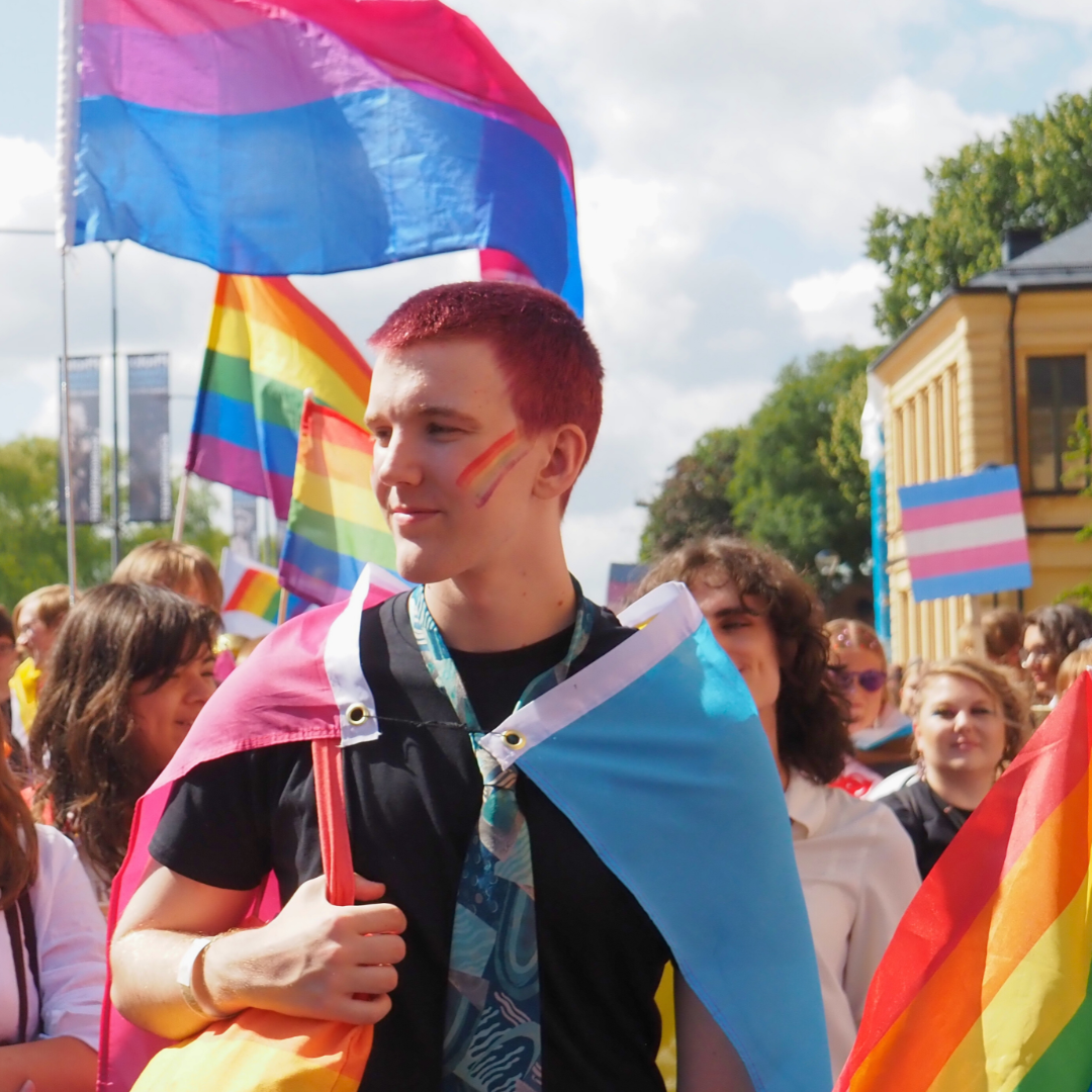 Foto på deltagare i vårt paradblock från Stockholm Pride 2023. En person är insvept i flaggan för pansexuella och har en sminkning och tygkassa med regnbågsflaggan. Den har svarta kläder och en mönstrad slips.