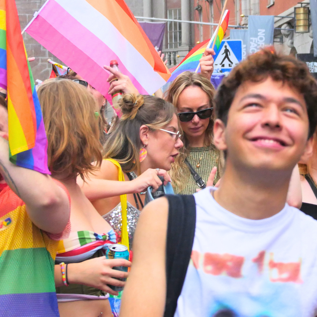 Foto på deltagare i Stockholm Pride parade 2024. Vissa är glada och fotar medan andra gör grimaser och pekar finger. Många viftar med regnbågsflaggor och flaggan för lesbiska.