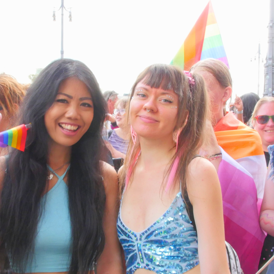 Foto på deltagare i vårt paradblock från Stockholm Pride 2024. Två personer håller hand och ler mot kameran: den ena håller i en liten regnbågsflagga och gör peace-tecknet.