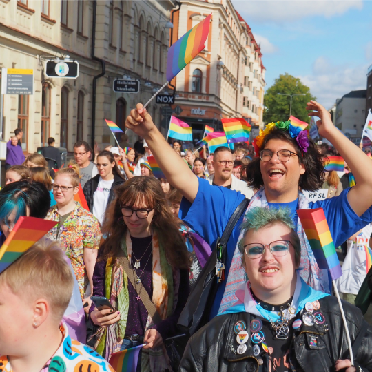 Foto på deltagare i paraden från Uppsala Pride 2023. De flesta viftar med regbågsflaggor och flaggan för pansexuella. De flesta ser antingen glada eller allvarliga ut.