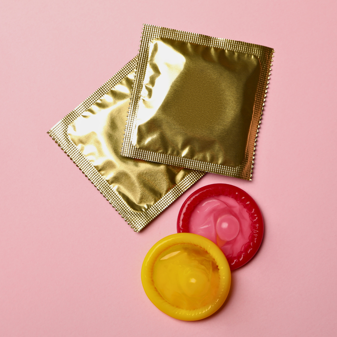 Närbild på två guldfärgade kondomförpackningar, en gul kondom och en röd kondom mot en rosa bakgrund.