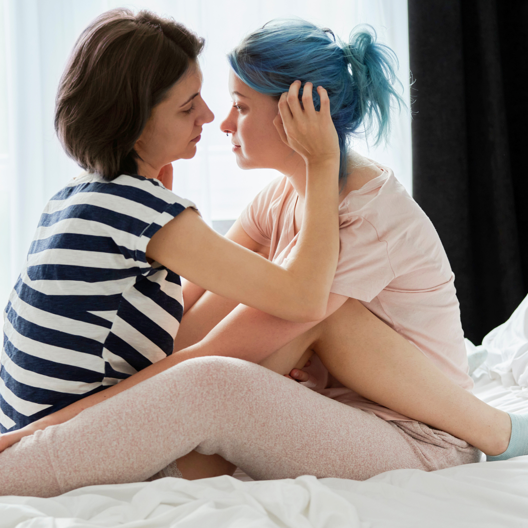 Foto på två personer som sitter i en säng och håller om varandra. Personerna har olika frisyrer, accessoarer och kläder.