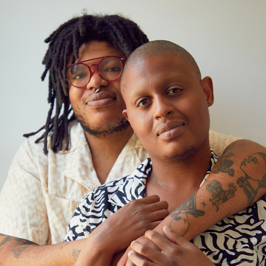 Foto på två personer som håller om varandra mot en grå bakgrund. Personerna har olika frisyrer, accessoarer och kläder.