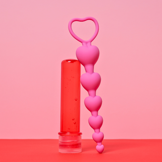 Foto på en röd flaska med glidmedel och rosa analkulor som är hjärtformade. Bakgrunden är rosa och röd.