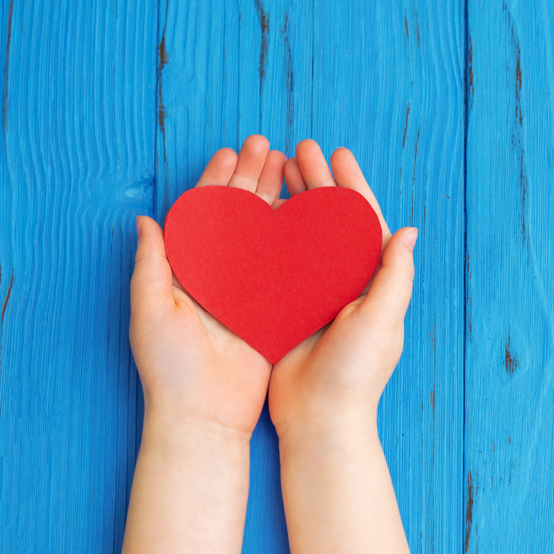 Närbild på två händer som håller upp ett rött pappershjärta framför en blå bakgrund.