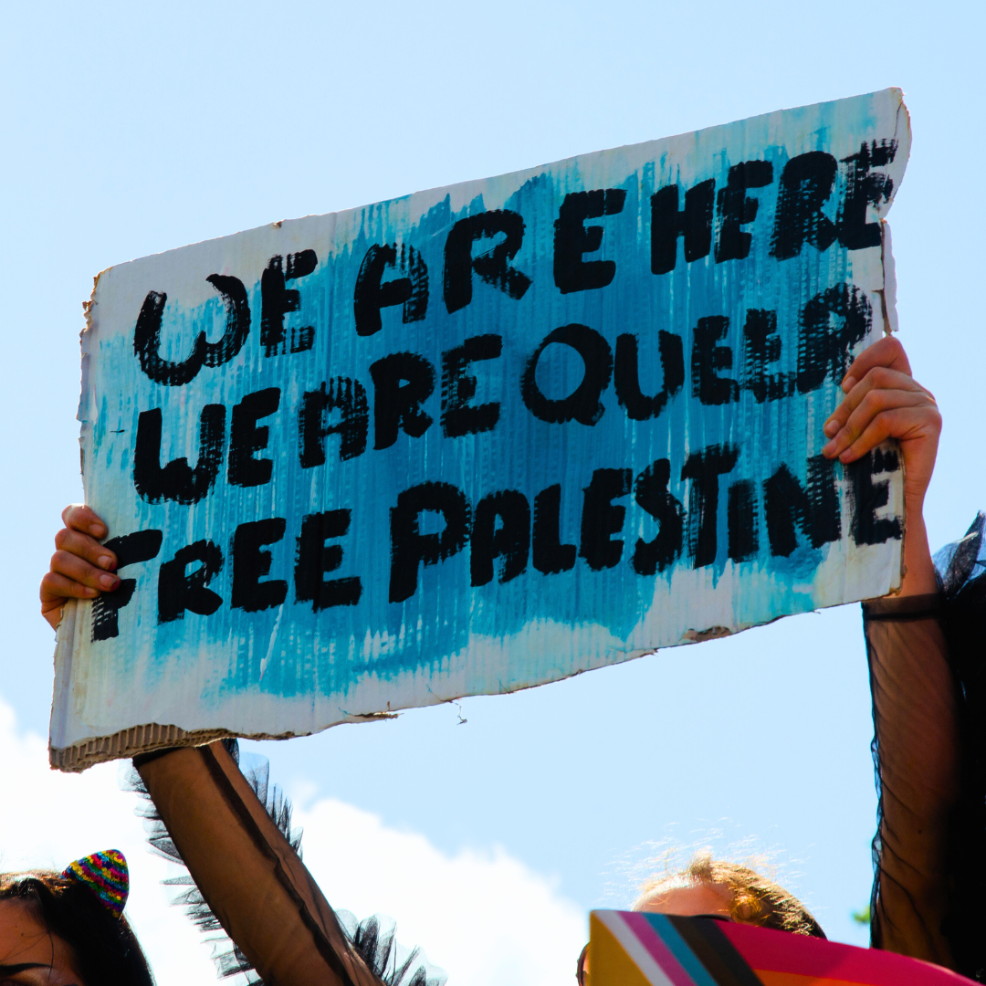 Foto på ett plakat som hålls upp mot en blå himmel. På plakatet står det "We are here, we are queer, free palestine" med svart text. I framgrund syns Progress pride-flaggan.