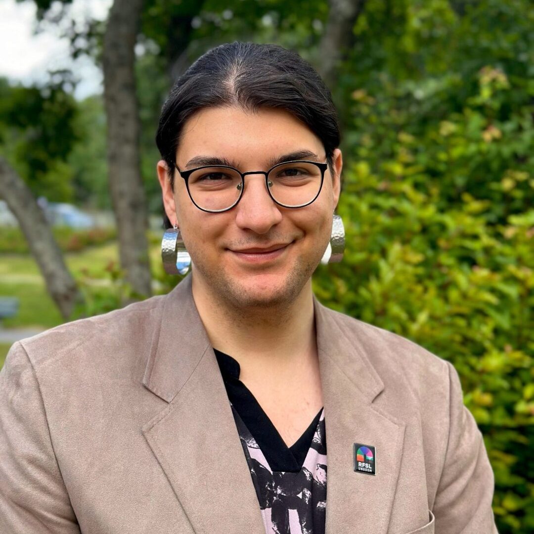 Bild på Elias Fjellander. Han har på sig svarta glasögon, silvriga örhängen, en svart topp och en grå kavaj med en RFSL Ungdom-pin.